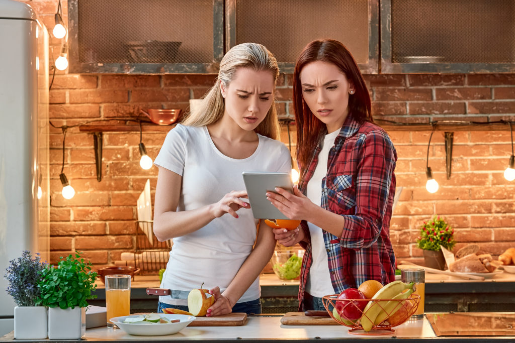 Portrait of two attractive women, having fun, while preparing fruit salad. They are fully involved in the process. Blonde girl in white T-shirt and her dark-haired friend in checkered shirt searching for healthy recipes, using tablet pc. They are impressed by variety of choice. Front view